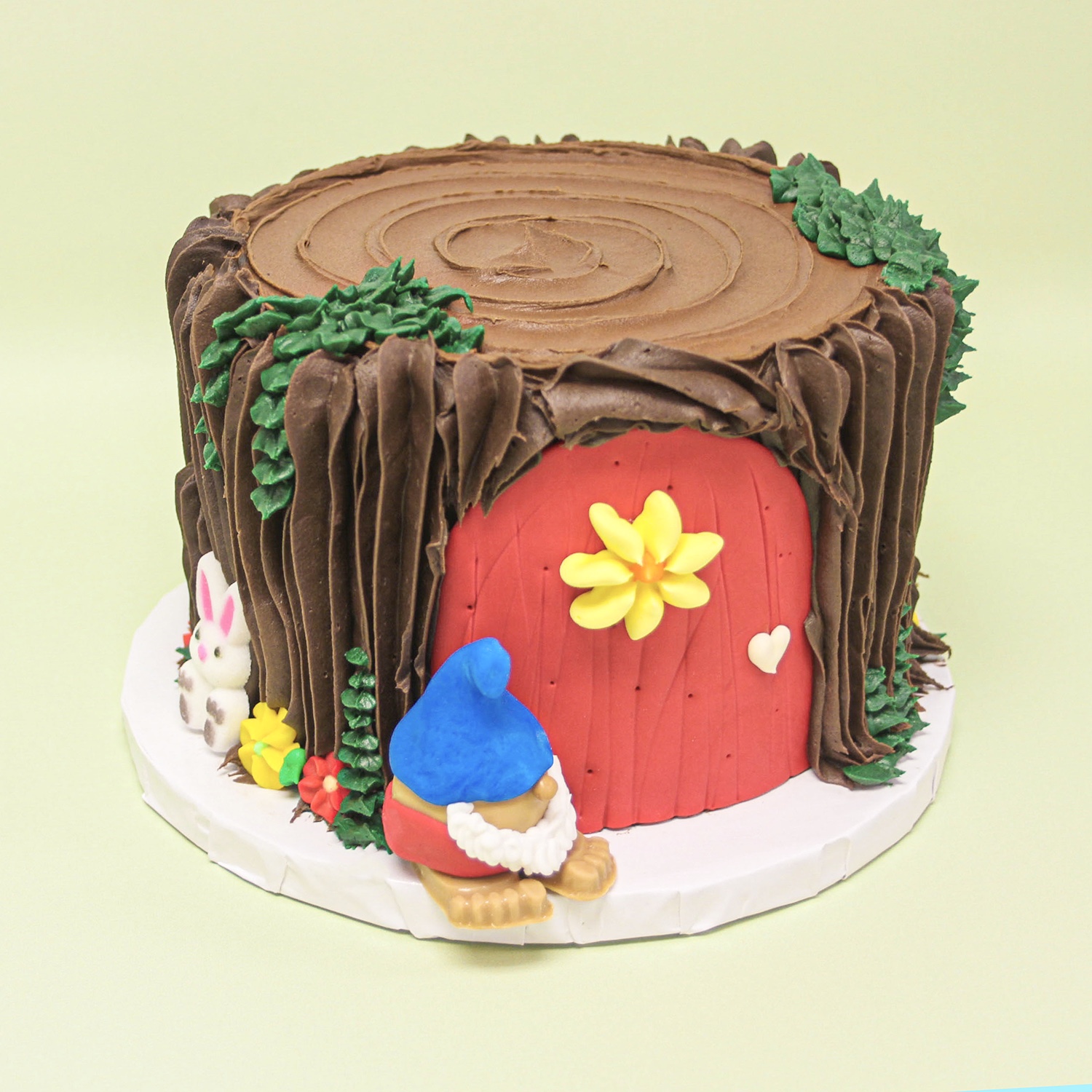 Gnome Tree Stump Cake with green buttercream leaves, chocolate buttercream bark, a gnome truffle, a red fondant door and icing sugar leaves and woodland animals.