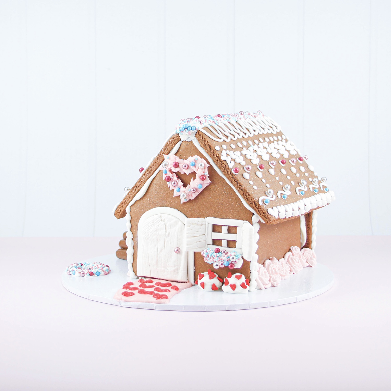 Front side right angle of valentine gingerbread house piped with royal icing accents of hearts , flowers, window trim and flowerbox, sidewalk of heart shaped sprinkles and a roof adorned with royal icing piping and sprinkles