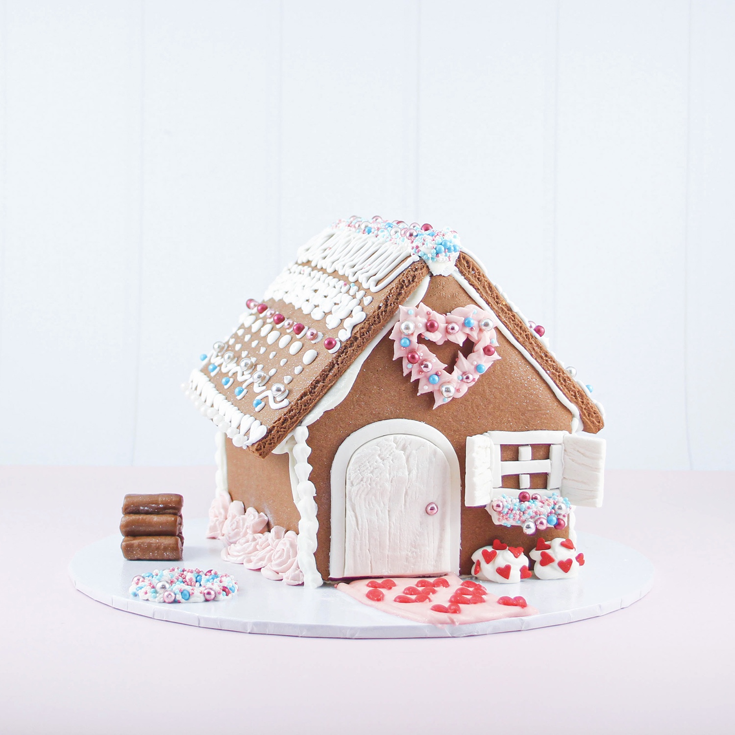 front and left side angle of valentine inspired gingerbread house, decorated with a fondant rose border flower bed, tootsie roll logs and hand piped accents and sprinkles