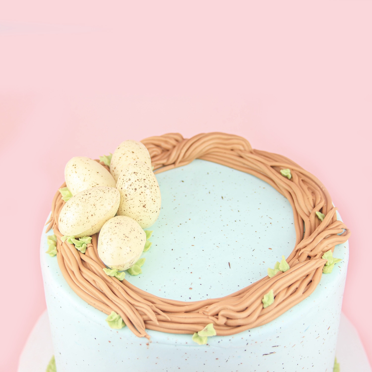 Top of cake with brown buttercream birdnest adorned with green piped leaves and handmand white chocolate speckled eggs