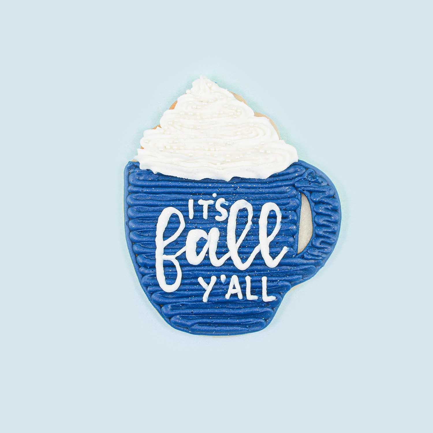 a blue mug piped in blue buttercream zig zags with freehand piping saying It's Fall y'all!  Piped whipped cream overflowing the top of the mug.