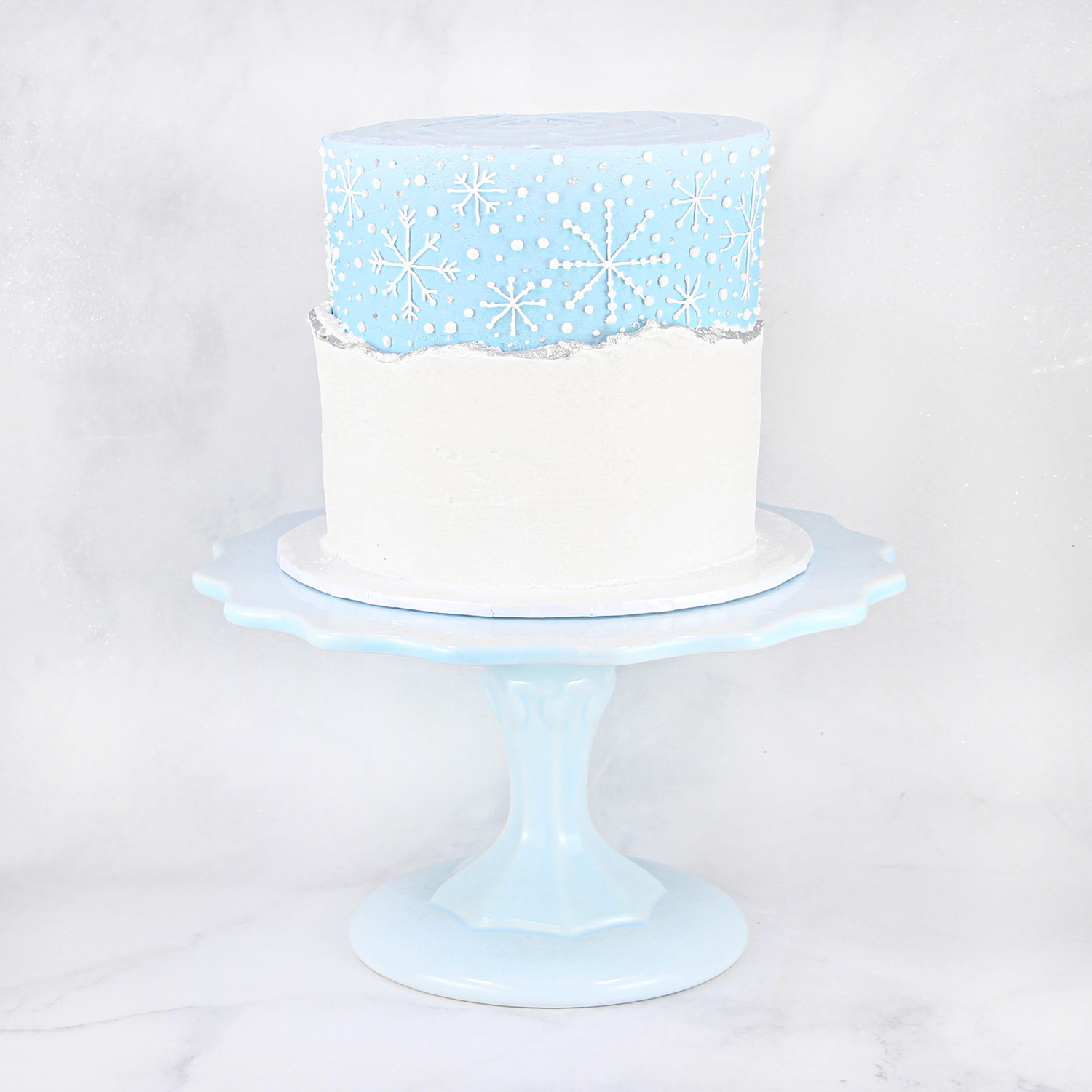2 layer cake covered in blue buttercream with faultline snow live edge painted in silver luster dust and hand piped snowflakes.