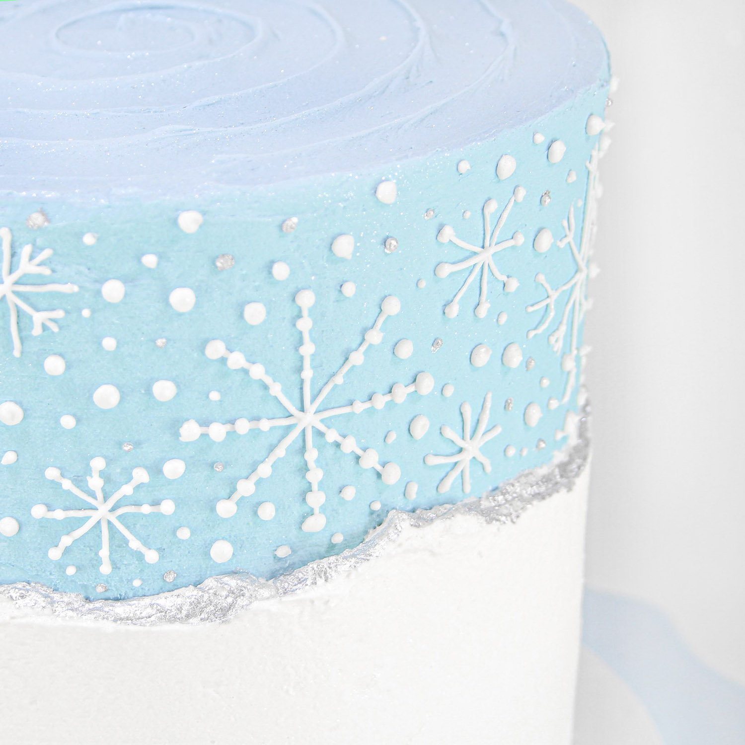 Hand piped snowflakes on blue buttercream just above a live edge snow faultline border painted in silver luster dust.