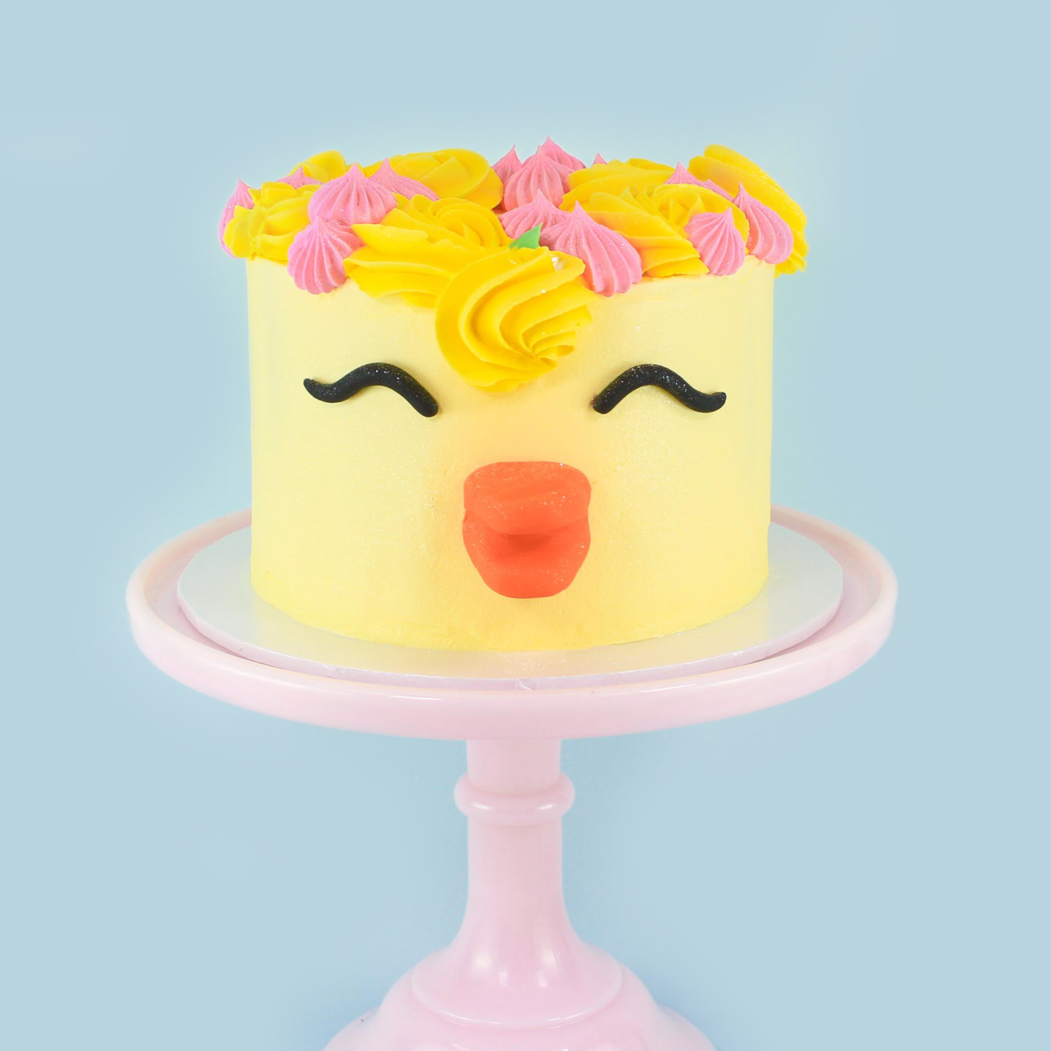 Cake with smooth yellow buttercream, adorned with yellow piped rosettes and pink piped dollops and decorated green buttercream eaves.  Black fondant rolled eyes and yellow fondant beak.  The floral crown is sprayed with edible jewel glitter dust