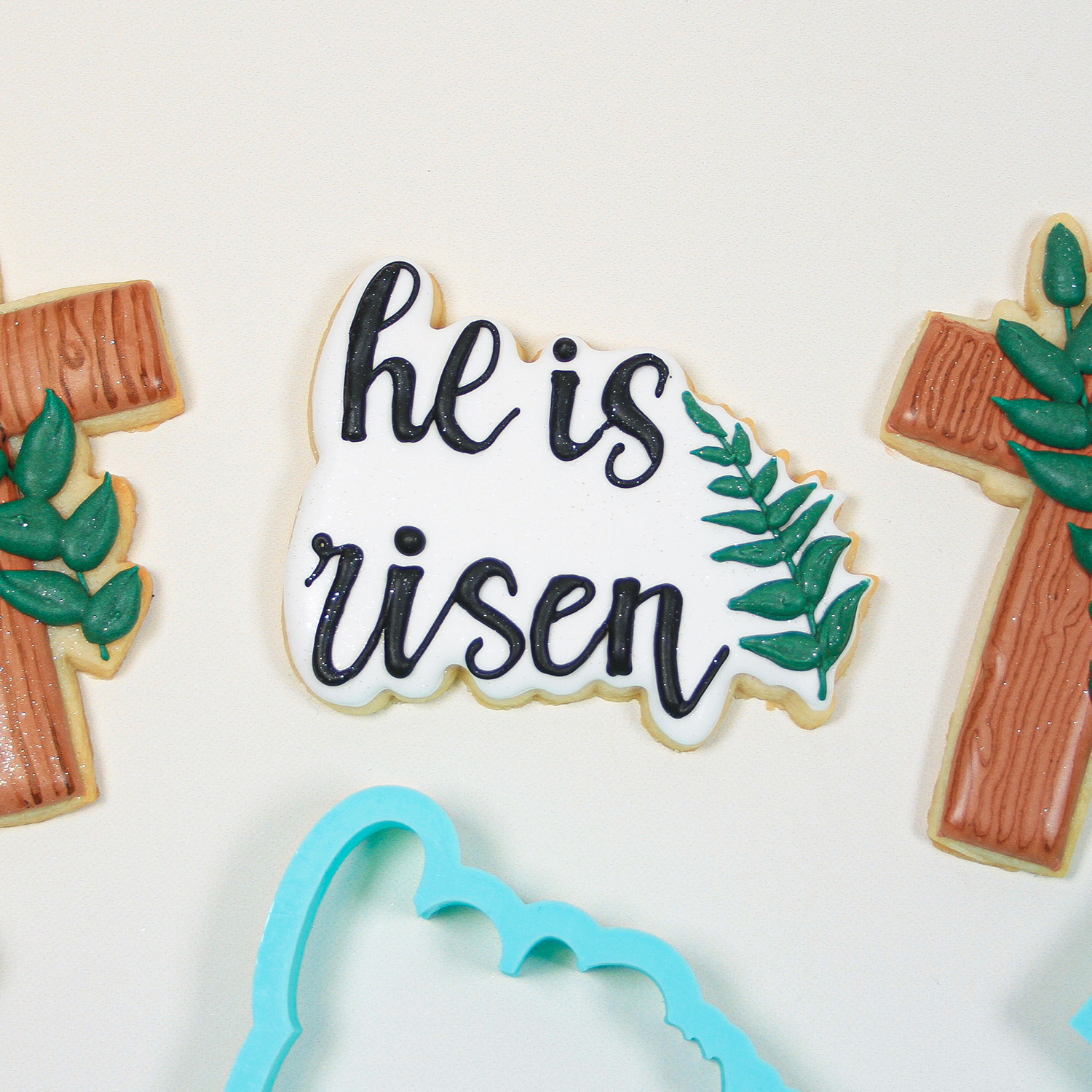 He is Risien Royal Icing decorted cookie with He is risen writing in black icing.  Cookie is adorned with green icing stem and leaves.
