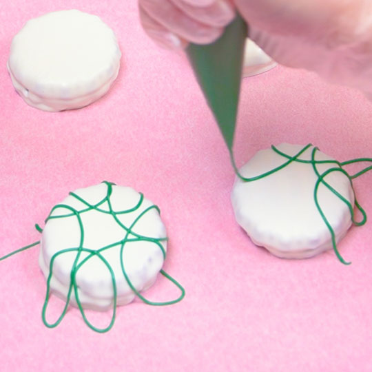 piping melted green chocolate onto chocolate dipped sandwich cookie